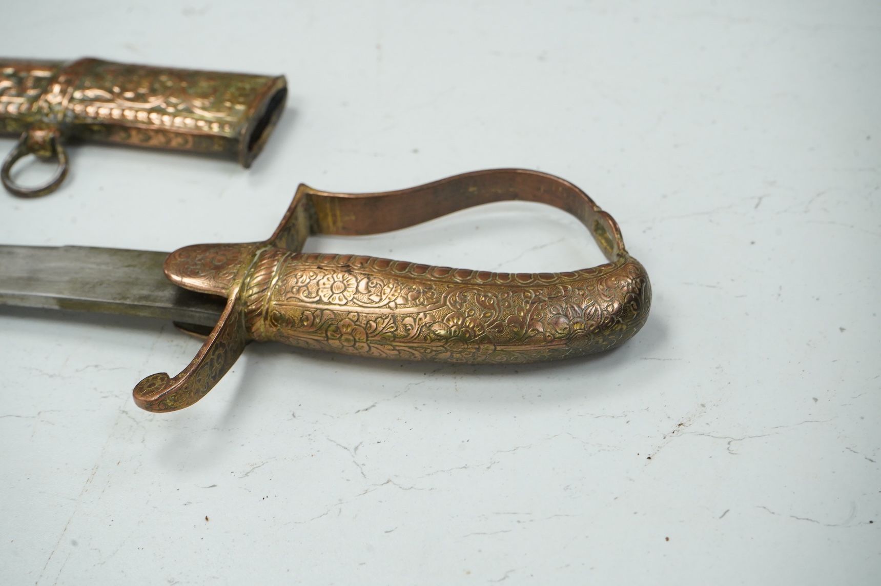 An early 19th century Indian sabre with curved single edge blade, copper grip and stirrup hilt, in its copper scabbard embossed with lions, scrolling foliage and flowers